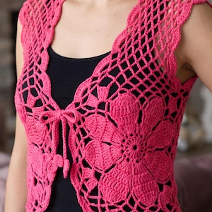May include: A bright pink crochet vest featuring an intricate openwork pattern with large floral motifs. The garment has a sleeveless design, a V-neckline, and a thin matching crochet tie closure at the center front, worn over a black top.