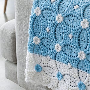 Puede incluir: Una manta de ganchillo con un patrón floral repetitivo en azul claro y blanco. El cuerpo principal es azul claro con pequeños motivos de flores blancas, mientras que el borde inferior es blanco con pequeños motivos de flores azul claro y un borde festoneado.