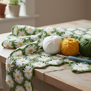 May include: A crochet blanket featuring a repeating pattern of white daisy motifs with yellow centers on a green background. Three yarn balls in white, yellow, and green sit on the blanket alongside a blue-handled crochet hook.
