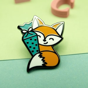 Könnte beinhalten: Orangefarbener und weißer Fuchsemaille-Pin, der eine türkisfarbene und schwarz gepunktete Geschenktüte hält. Der Pin befindet sich auf einem grün-gelben Hintergrund.