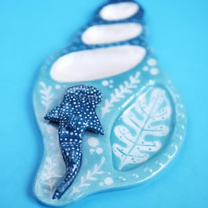May include: A ceramic dish shaped like a seashell, glazed in light blue with white painted details. It features a three-dimensional whale shark figure on the surface, alongside white painted leaf patterns and small circular accents.