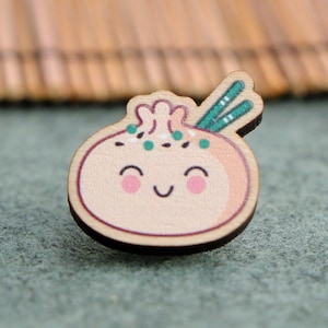 Könnte beinhalten: Eine Holz-Anstecknadel in Form eines lächelnden Knödels mit grünen Akzenten und zwei Essstäbchen oben drauf.