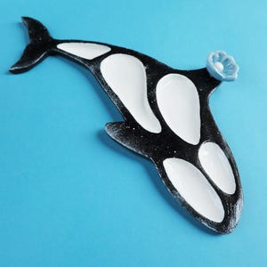 Puede incluir: Una bandeja de servir con forma de orca en blanco y negro con cuatro secciones blancas hundidas distintas. El cuerpo es negro con un acabado texturizado y un pequeño adorno en forma de flor azul claro está fijado cerca de la parte superior.
