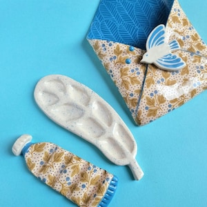 Pode incluir: Um conjunto de cerâmica composto por um bolso de parede em forma de envelope com um pássaro azul, uma paleta de pintura em forma de folha com manchas azuis e um tubo de tinta. O padrão apresenta folhas castanhas e bagas azuis sobre fundo creme.