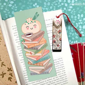 Könnte beinhalten: Ein grünes Lesezeichen mit einer Cartoon-Illustration eines Bao-Buns. Der Bao-Bun lächelt und hat eine kleine Schüssel Suppe oben drauf. Das Lesezeichen zeigt einen Stapel Bücher mit Titeln wie "Dim Sum", "Bao Bao Bao", "Soul Food", "Rezepte", "Foodie Stories" und "Süße Erinnerungen".