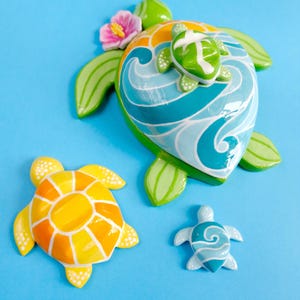 May include: A large sea turtle figurine with a blue and white wave pattern on its shell, topped with a small green turtle and a pink hibiscus flower. Beside it are a smaller yellow and orange turtle figurine and a tiny blue and white turtle.