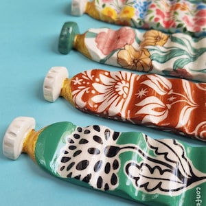 Può includere: Una serie di tubetti di vernice in ceramica disposti in fila, ognuno caratterizzato da distinti motivi floreali e botanici dipinti a mano in vari colori, tra cui verde, arancione e bianco, con il testo confetti.friends visibile in basso.