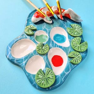 Może przedstawiać: Ceramiczna paleta malarska w kształcie stawu z biało-pomarańczową rybą koi na górze. Paleta posiada kilka białych wgłębień na farbę, niektóre zawierają niebieski, zielony i czerwony pigment, otoczone zielonymi liśćmi lilii wodnych.