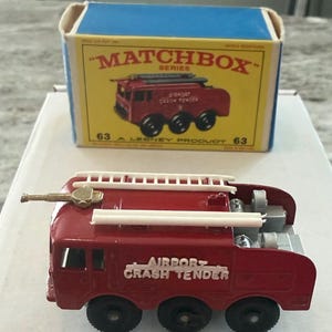 May include: A vintage Matchbox Series toy fire truck, model 63, in red with white accents. The toy reads "AIRPORT CRASH TENDER" and includes a ladder and a water cannon. The box is blue and yellow with the Matchbox logo.
