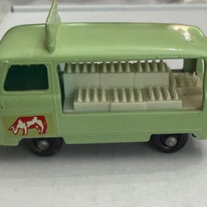May include: A light green toy ice cream van with a red and white cow logo on the side. The van has a white interior with rows of ice cream cones. The van has black wheels and a fin on top.