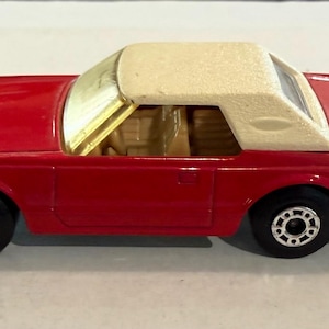 Puede incluir: Un coche de juguete rojo con techo color crema. El vehículo en miniatura tiene ruedas negras con llantas plateadas y un interior beige. El coche es un modelo clásico, probablemente de mediados del siglo XX.
