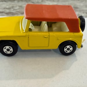 May include: A yellow toy car with a red top and black tires. The miniature vehicle has a spare tire mounted on the back and white interior details. The toy car is a vintage collectible.