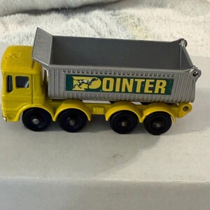 May include: A yellow toy dump truck with a gray bed. The truck has black wheels and the word "POINTER" in green and yellow on the side. The truck is sitting on a white surface.