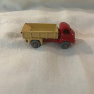 May include: A red toy dump truck with a tan bed. The truck has four wheels and a cab with a windshield.