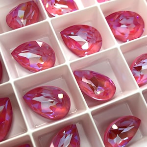May include: A close-up of a pink iridescent pear-shaped rhinestones in a white plastic storage container.