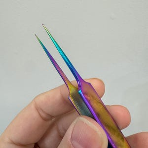 May include: A pair of iridescent tweezers with a rainbow finish. The tweezers have a pointed tip and are held in a hand. The colors shift from gold to purple, blue, and green.