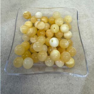 May include: A clear, square dish filled with numerous yellow and off-white round beads. The beads have a smooth, polished appearance and are evenly distributed within the dish. Each bead has a small hole, suggesting they are for stringing.