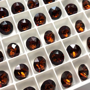 May include: A close-up view of a white plastic tray filled with round, faceted, dark brown glass rhinestones. The rhinestones are arranged in rows and columns.