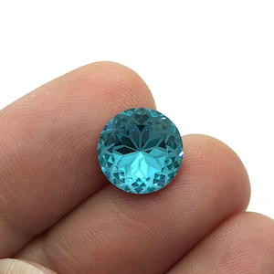 May include: A round, faceted gemstone in a vibrant turquoise hue. The gem has a complex pattern of cuts that reflect light, creating a sparkling effect. The gemstone is held in a person's hand.