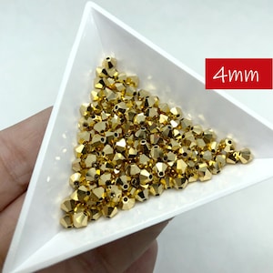 May include: A collection of small, faceted gold beads, approximately 4mm in size, are displayed in a white triangular tray. The beads have a metallic sheen and reflect light. The text "4mm" is in a red rectangle.