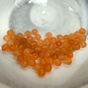 May include: A collection of small, round, orange beads. The beads are scattered in a white bowl. The beads have a translucent quality, and the color varies from light to dark orange. The bowl has a marbled appearance.