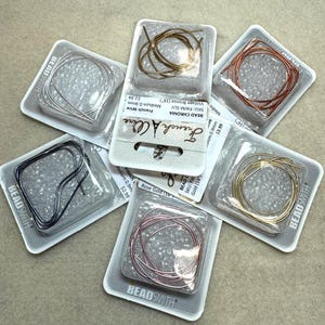 May include: Assortment of BeadSmith French wire in various colors, including silver, gold, rose gold, and bronze. Each wire is packaged in a clear plastic blister pack. The packages are labeled with the BeadSmith logo and product information.