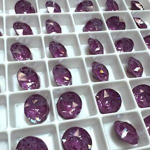 May include: A close-up view of a white plastic tray filled with small, round, purple iridescent rhinestones. The rhinestones are arranged in rows and columns.