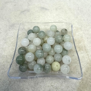 May include: A collection of round, translucent beads in shades of green and white, displayed in a clear, square dish. The beads vary in color intensity, creating a natural, organic aesthetic. The beads are likely used for jewelry making or crafting.