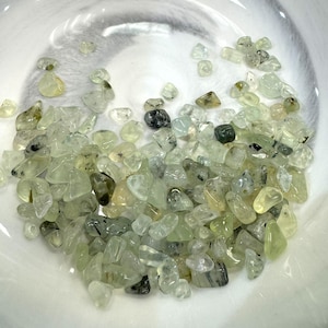 May include: A collection of small, tumbled prehnite gemstones in shades of pale green, yellow-green, and clear. The stones are scattered in a white, shallow bowl, showcasing their translucent quality and natural variations.