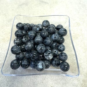 May include: A clear square dish filled with numerous dark gray, round beads. The beads have a glossy finish and appear to be made of a stone-like material. Each bead has a small hole drilled through it, suggesting they are for stringing.