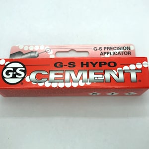 Könnte beinhalten: Eine rote Tube G-S Hypo Cement mit einem weißen Etikett und einem weißen Kreis mit den Buchstaben G-S darin. Das Etikett trägt die Aufschrift "G-S Hypo Cement" und "G-S Precision Applicator".