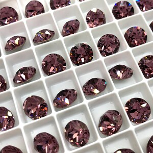 May include: A close-up view of a white tray filled with small, round, purple rhinestones. The rhinestones are arranged in rows and columns, and they have a sparkly, iridescent finish.