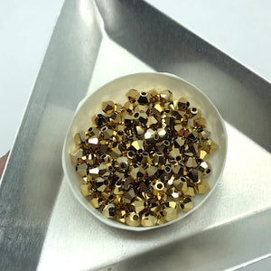 May include: A white bowl filled with gold-colored faceted glass beads. The beads are in a triangular shape and are arranged in a random pattern.
