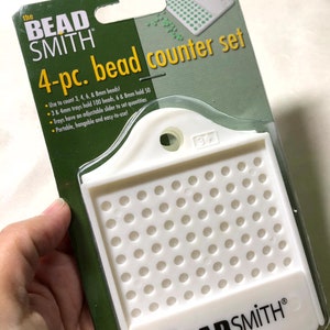 May include: A white plastic bead counter set with four trays. The trays are labelled "The Bead Smith" and have a hole for hanging. The trays are designed to hold 3, 4, 6, and 8 mm beads.
