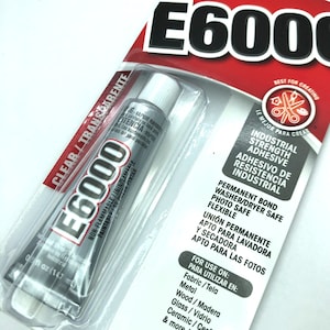 May include: E6000 industrial strength adhesive in a clear tube, packaged in a blister pack. The label includes the text "CLEAR/TRANSPARENTE" and "NON-FLAMMABLE/ININFLAMABLE". The tube contains 0.5 fl oz (14.7 mL) of adhesive.