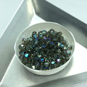 May include: A close-up of a small bowl filled with iridescent green glass beads. The beads are faceted and have a shimmery finish.