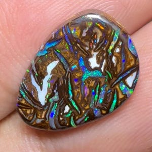 Puede incluir: Una piedra preciosa de ópalo pulida, de forma ovalada. La piedra presenta un color base marrón oscuro con intrincados patrones de iridiscencia blanca, azul, verde y dorada. La superficie del ópalo es lisa y reflectante.