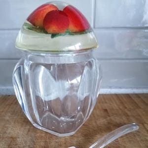 May include: A clear glass jar with a decorative lid featuring three red and orange fruit. A clear plastic spoon rests beside the jar on a wooden surface. The jar has a faceted design.