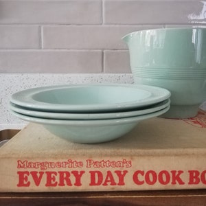 May include: Three light blue bowls stacked on a vintage cookbook, alongside a matching pitcher. The bowls and pitcher are a pale turquoise colour. The background is a light grey tiled wall.