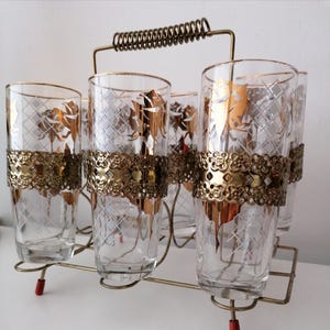 May include: A set of six clear drinking glasses with gold floral patterns and decorative gold bands. The glasses are held in a gold metal carrier with a handle and red feet.