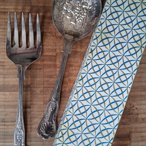 May include: A silver-toned metal serving fork and a matching serving spoon with an embossed floral and fruit pattern on the bowl, accompanied by a rectangular box featuring a repeating blue and gold geometric circle pattern on its exterior.