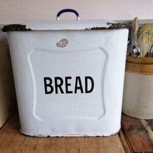 May include: A white enamel bread bin with a blue handle and the word "BREAD" in black. The rectangular container shows signs of wear, with a distressed finish. It is likely a vintage kitchen item.