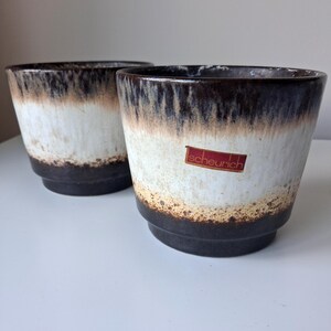 May include: Two ceramic plant pots featuring a dark brown base and rim with a white centre section. The glaze transitions from dark brown to a lighter cream tone. A red rectangular sticker with the white text scheurich is on the front pot.