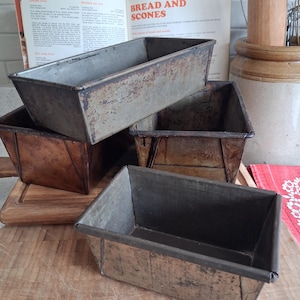 May include: Several vintage metal loaf tins, showing signs of age and use. The tins are arranged on a wooden chopping board, with an open cookbook titled "Bread and Scones" in the background. The tins are various sizes and shapes, perfect for baking.