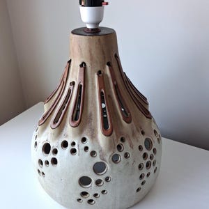 May include: A ceramic lamp base with a bulb socket at the top. The body features a beige and brown gradient finish with vertical elongated cutouts near the neck and various circular perforations scattered across the lower rounded section.
