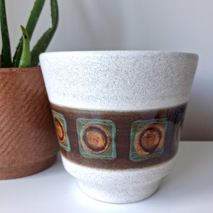 May include: A ceramic planter with a speckled off-white finish on the top and bottom sections. The centre features a wide, dark brown horizontal band decorated with a repeating pattern of teal square outlines surrounding circular orange centres.