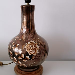 May include: A decorative table lamp with a brown metallic finish and floral design. The lamp features a bulbous body with hand-painted flowers in cream and brown. It sits on a wooden base and has a black lamp socket.