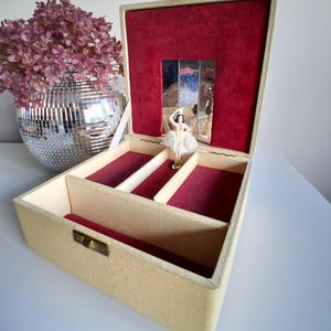 May include: A cream-coloured jewellery box with a hinged lid, featuring a red velvet interior lining, a small rectangular mirror, and a miniature ballerina figurine wearing a white lace tutu that stands in the centre of the open storage compartment.