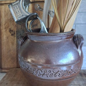 May include: A brown ceramic utensil pot with a floral design. It holds kitchen tools such as a metal egg beater, wooden spoons, and a whisk. The pot has two handles and is on a wooden surface. The pot is approximately 20 cm in height.