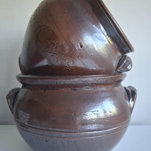 May include: A brown ceramic pot with a lid. The pot has two handles and a decorative band around the middle. The lid is slightly ajar and has a decorative pattern. The pot is likely for cooking or storage.